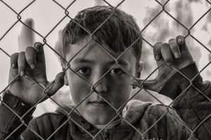 Photo en noir et blanc d'un jeune garçon blanc regardant l'objectif derrière une barrière quadrillée. Ses mains sont posées sur la barrière. Il porte un gilet, il y a des yeux noirs et des cheveux noirs.