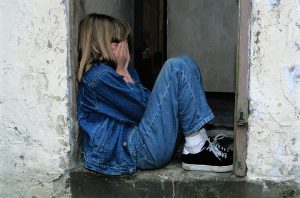 Photo prise de profil d'une jeune fille blanche assise sur le palier d'une porte. Elle porte une veste et un pantalon en jeans. Elle porte des chaussures noires. Ses deux mains sont sur son visage.