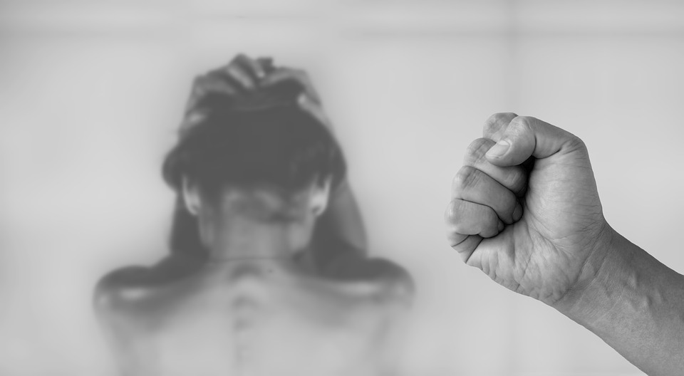 Photo en noir et blanc d'une main avec le poing serré qui se trouve sur la partie droite de la photo. En arrière plan, une femme blanche portant des cheveux noirs, de dos, est torse nu, assise et se tient le visage pour se protéger.