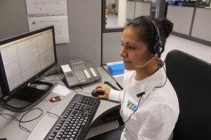 Photo d'une femme noire portant un casque avec un micro sur la tête. Elle porte une blouse blanche. Elle regarde en direction d'un écran. Elle tient une souris d'ordinateur. Sur son bureau, un téléphone fixe, un clavier d'ordinateur et un écran d'ordinateur sur lequel est affiché des graphiques.