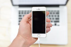 Photo d'une main tenant un smartphone de la main gauche. Le smartphone est branché à son chargeur. En arrière plan, un ordinateur ouvert posé sur une table en bois.
