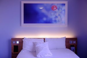 Photo d'une chambre d'hotel. Il y a un lien avec trois oreillers et des draps blancs. Sur chaque coté du lit, des interrupteurs. Au-dessus du lit, un cadre de couleur bleu.