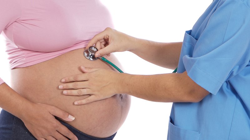 Photo d'une femme enceinte, sur la gauche de la photo, portant un t-shirt rose retroussé jusqu'à sa poitrine. Une dame utilise un stéthoscope avec la main droite et a la main gauche posé sur le ventre. Elle porte une blouse bleue.