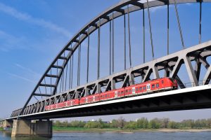 Photo d'un pont composé d'un arc gris par dessus. Au milieu de la photo, un train composé de 3 wagons de couleur rouge passe sur ce pont. Sous ce pont, de l'eau. En arrière plan, des arbres et la rive.