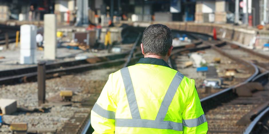 Photo prise de dos d'un homme portant une veste fluorescente jaune. En arrière plan, des lignes de chemin de fer en travaux.