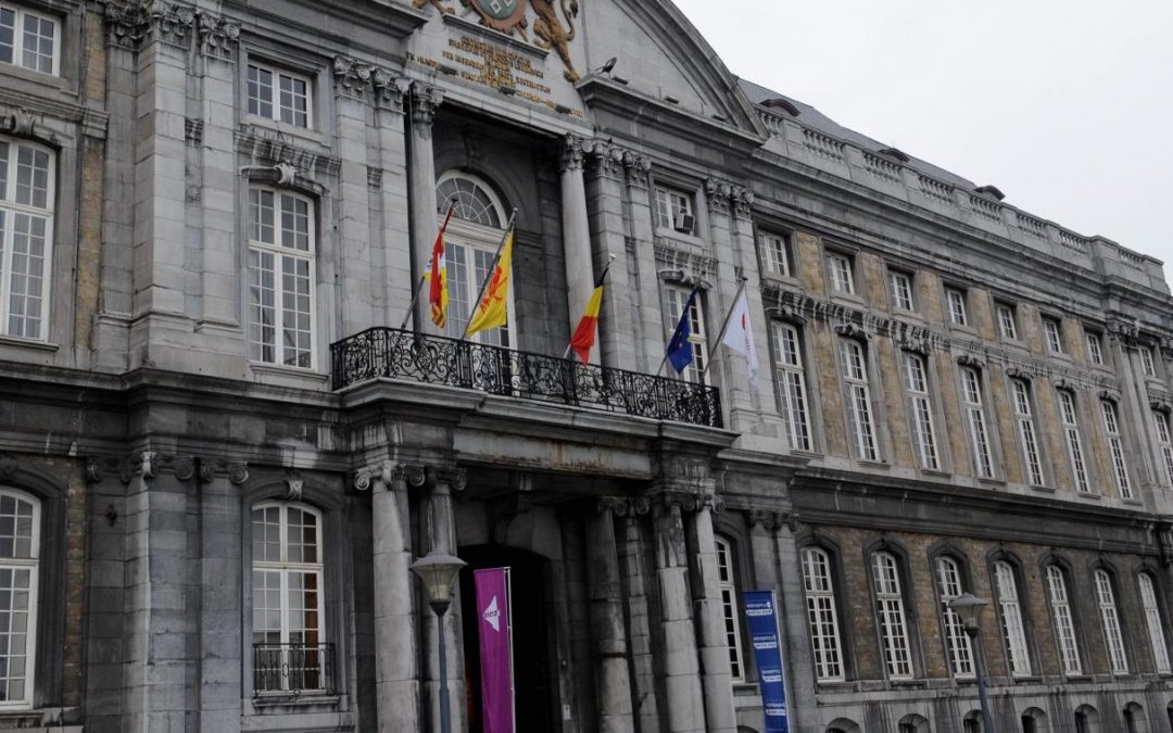 Photo de l'entrée du Palais de Justice de la Ville de Liège. L'entrée est ornée de piliers en brique grise. Sur le balcon du batiment, il y a 5 drapeaux: en partant de la gauche, le drapeau de la province de Liège, le drapeau de la Région Wallonne, le drapeau de la Belgique, le drapeau de l'Union Européenne et le drapeau de la communauté française