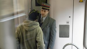 Photo prise dans un train où un homme que l'on aperçoit de face et portant un chapeau gris de la SNCB ainsi qu'une veste grise sous laquelle il a une cravatte et une chemise. En face de cet homme, et de dos sur la photo, un homme portant une casquette est face au controleur.