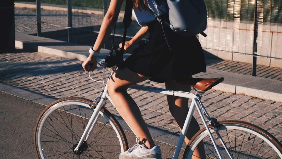 Photo d’une personne sur un vélo. Cette personne, dont on ne voit pas le visage, porte un sac à dos, un appareil photo est suspendu à son cou. Elle porte également un short noir et des baskets blanches.