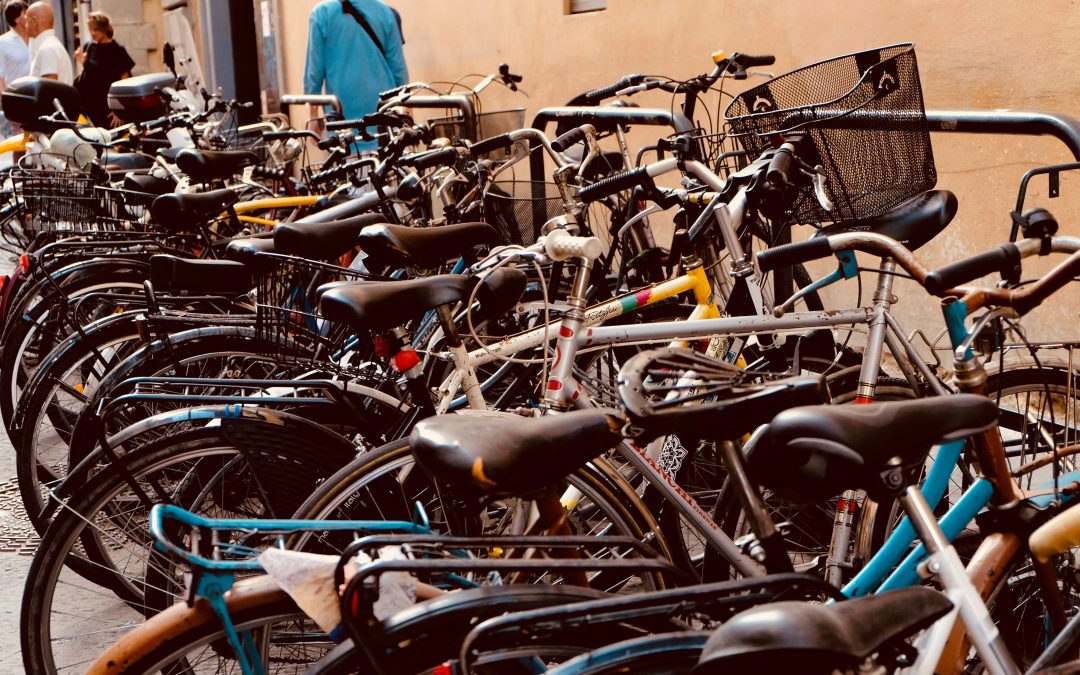Photo d’un parking pour vélo sur lequel se trouve un grand nombre de vélos aligné dans la même direction. Ce parking est situé au bord d’un trottoir sur lequel on aperçoit des personnes marchées.