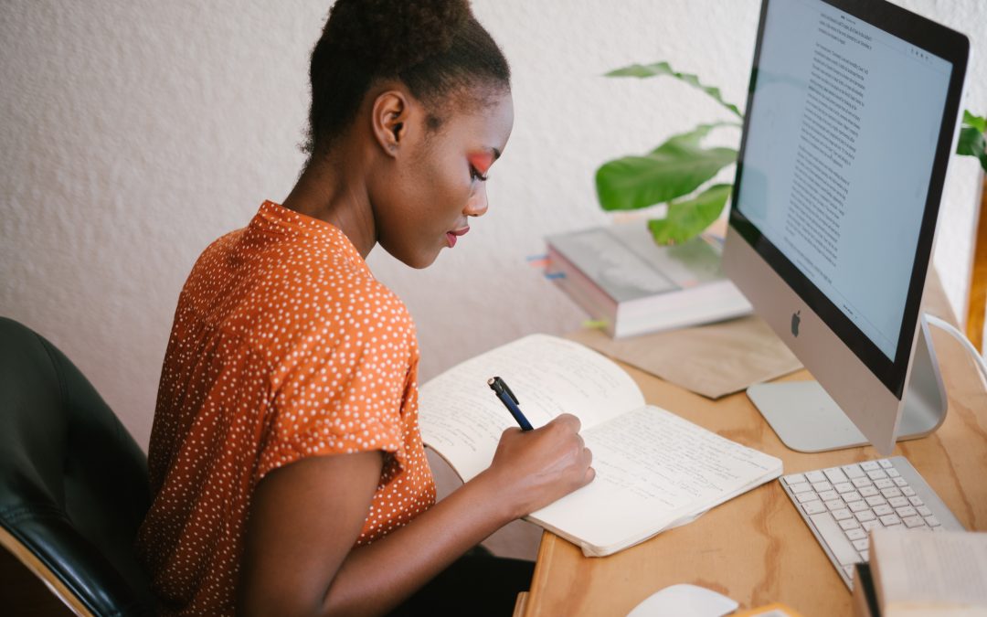 Une femme noire écrit dans un cahier. En face d’elle, un ordinateur Mac. A sa gauche, un cahier posé sur une table en bois clair. Elle est assise sur une chaise de bureau noir et porte une chemise avec des pointillés blancs sur fond orange. Sur son tableau, on trouve un clavier, une souris d’ordinateur, une plante et des livres.