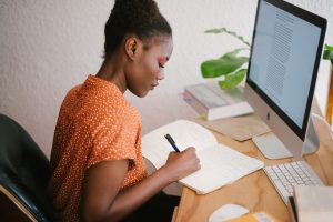 Une femme noire écrit dans un cahier. En face d’elle, un ordinateur Mac. A sa gauche, un cahier posé sur une table en bois clair. Elle est assise sur une chaise de bureau noir et porte une chemise avec des pointillés blancs sur fond orange. Sur son tableau, on trouve un clavier, une souris d’ordinateur, une plante et des livres.