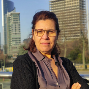 Hafida Bachir, Conseillère politique genre, sourit et regarde la caméra. Elle a les bras croisés. Elle porte une chemise mauve avec un gilet noir par-dessus. Elle porte une paire de lunettes. Elle se trouve à l'extérieur, face aux bâtiments du jardin botanique à Bruxelles.