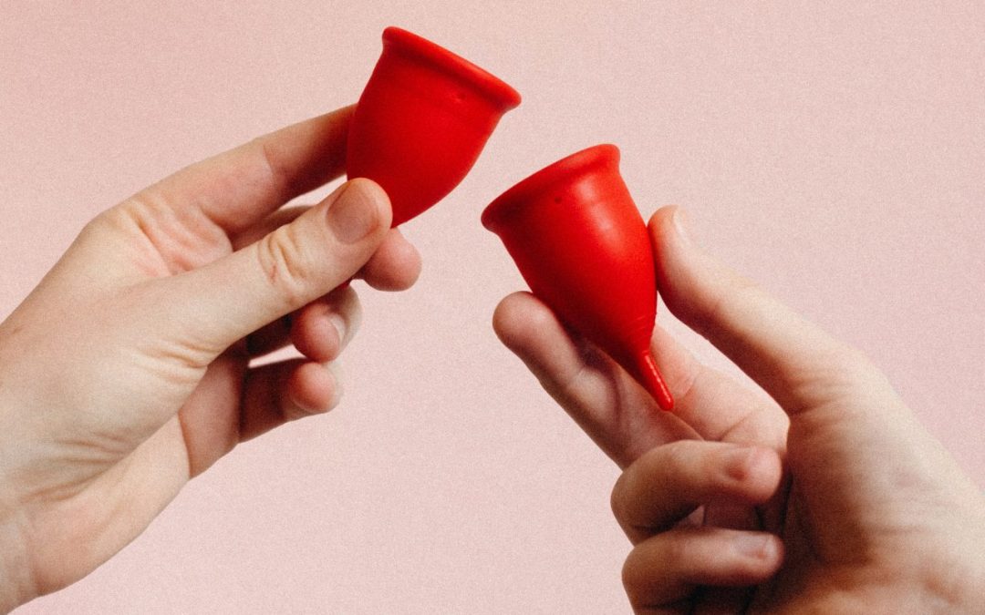 Sur un fond de couleur rose, deux mains tiennent deux coupelles menstruelles rouges.